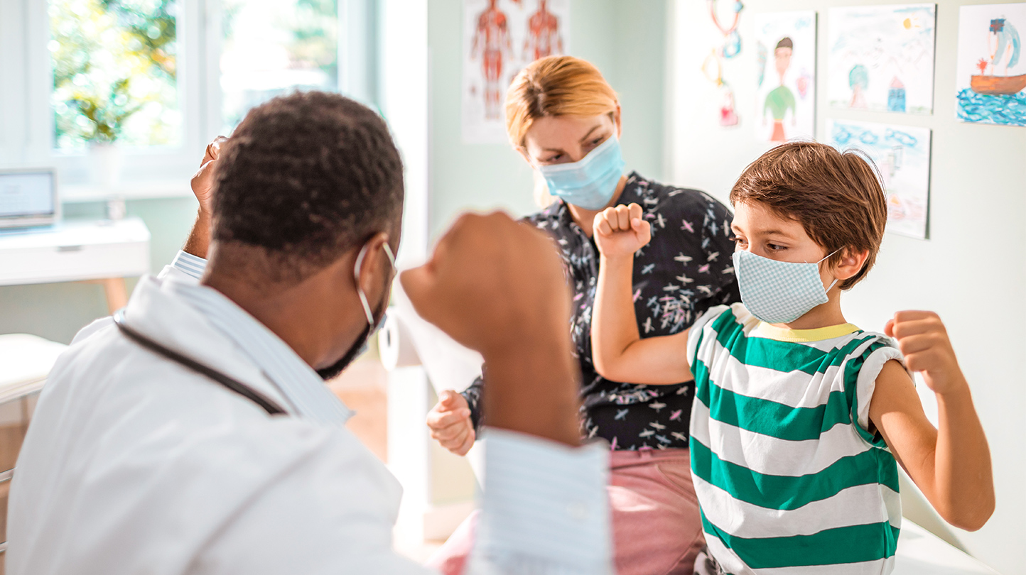Imagen de un médico, una madre y su hijo, todos con mascarillas en un consultorio, mientras el médico y el niño muestran juntos los músculos de sus brazos