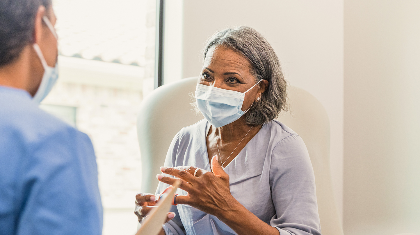 Image of a doctor and his patient wearing masks while she talks to him in the exam room