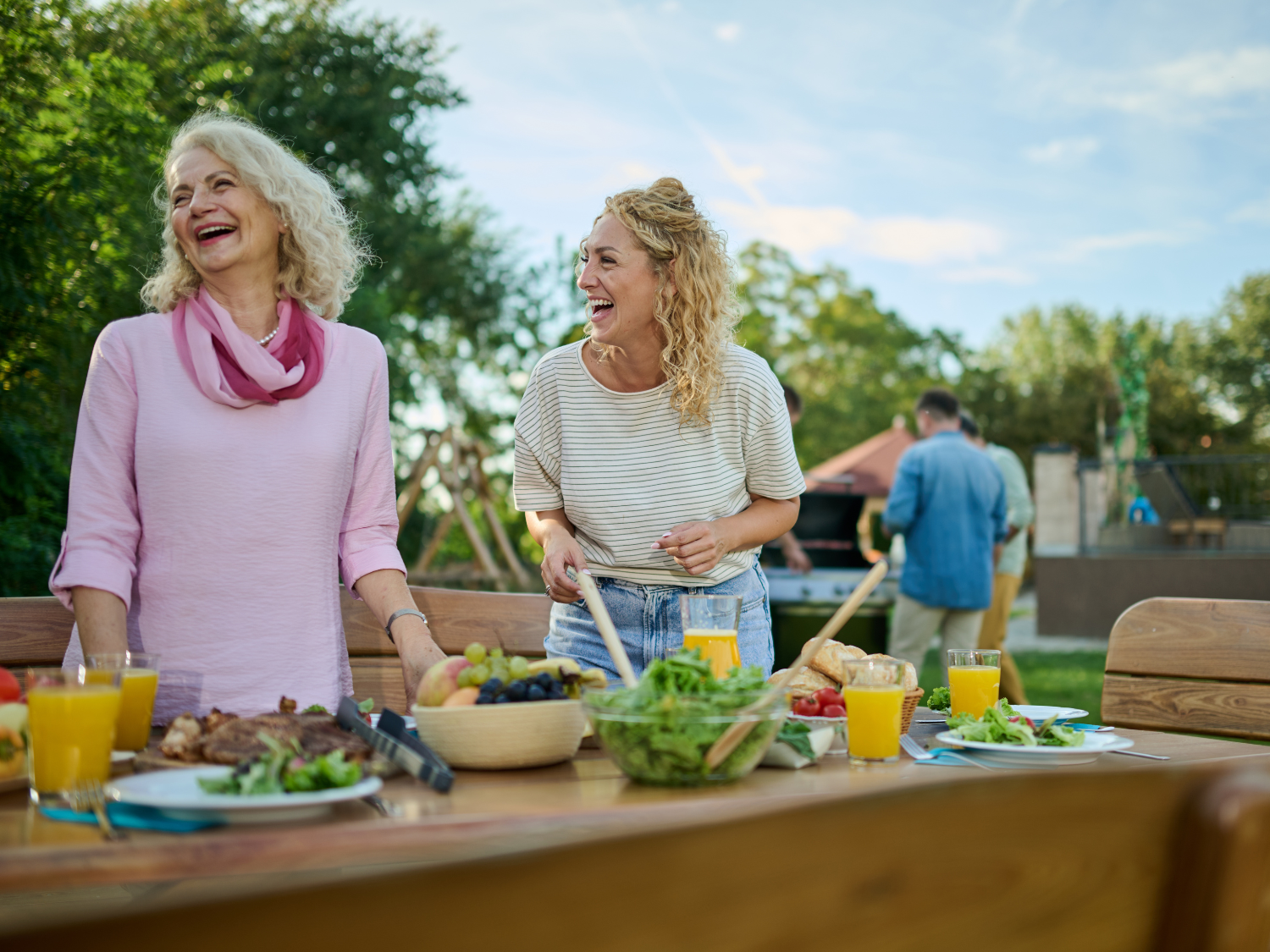 Two women laughing, standing over a table of food with men barbequing food behind them.