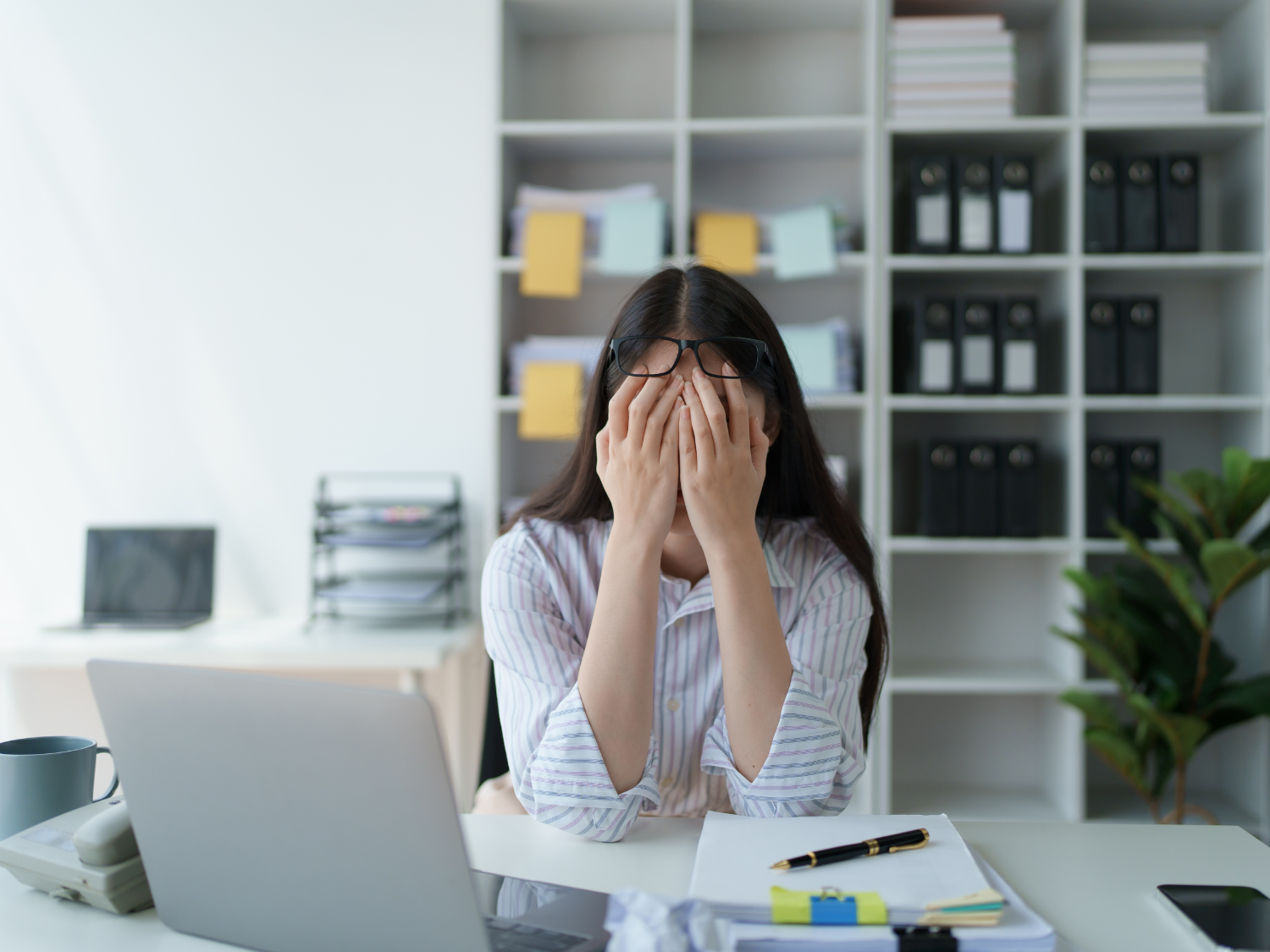 Woman sitting at desk in home office with hands over her face, showing symptoms of burnout.