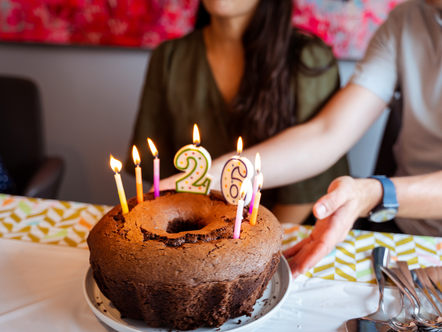 Close up of chocolate bundt cake with lit candles with the number 26 being placed infront of young adult female.