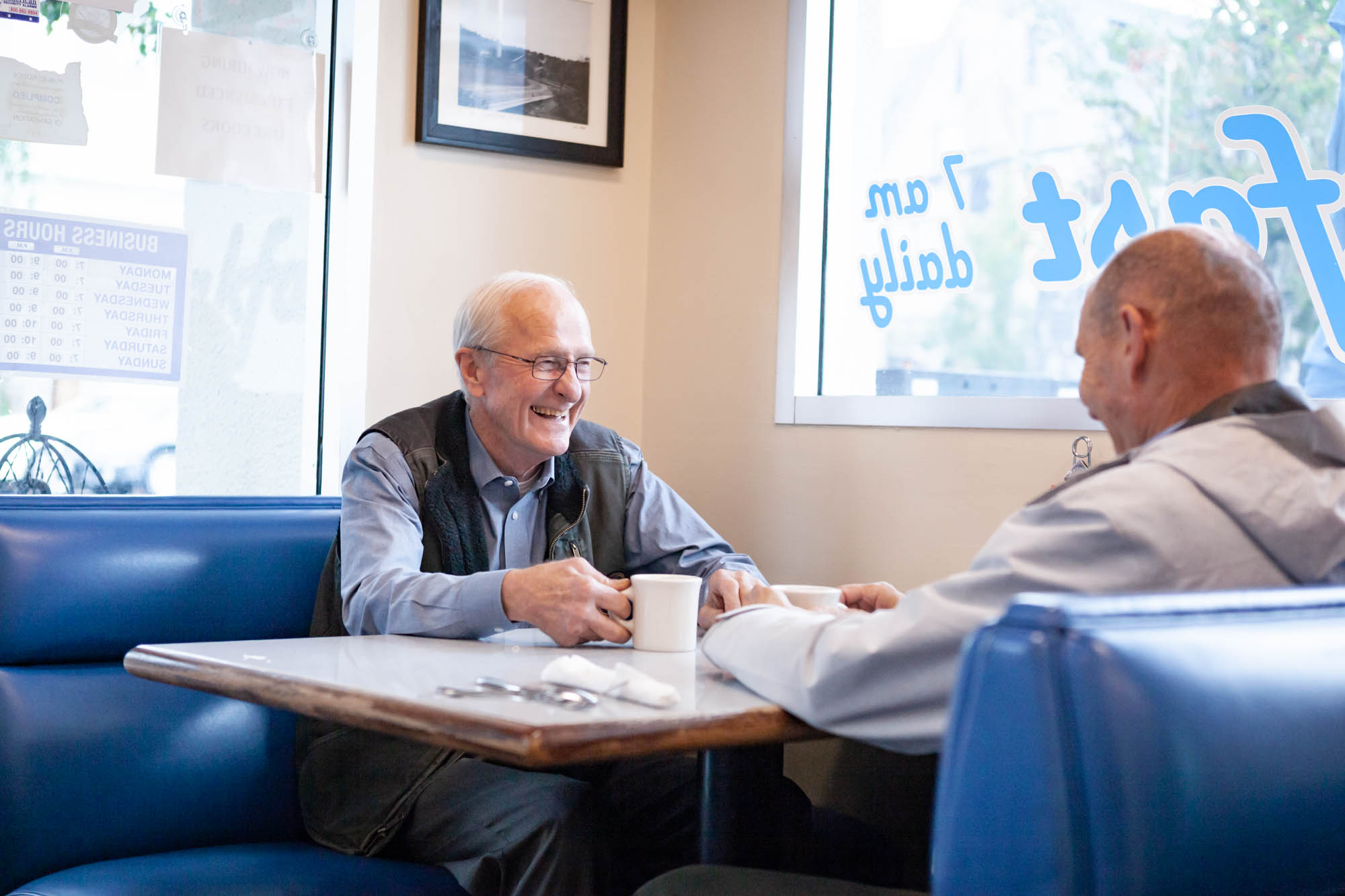 Two men sitting at a cafe table laughing and drinking coffee.